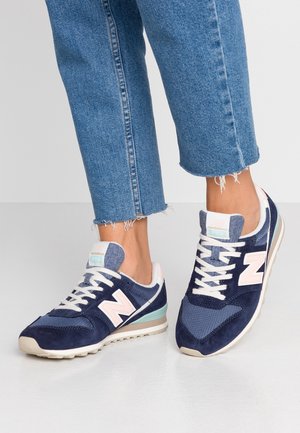 Person iført blå og hvide New Balance-sneakers og slidte blå jeans med flossede kanter står på en lys grå gulv.