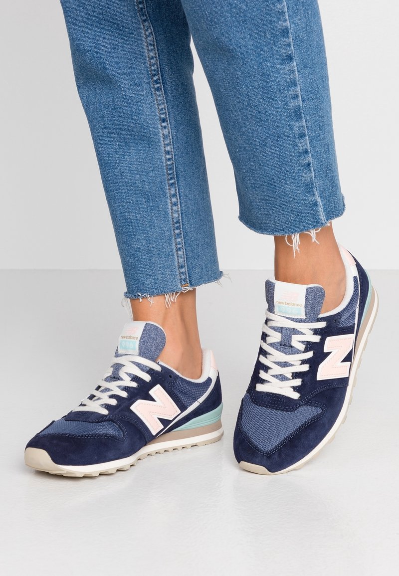 Personne portant des baskets New Balance bleues et blanches et un jean bleu à ourlet effiloché, debout sur un sol gris clair.