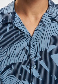 Camicia a maniche corte di colore azzurro chiaro con motivi lineari e a foglia blu scuro, dotata di colletto a punta e chiusura con bottoni.