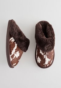 Brązowo-białe kapcie w wzór krowy z miękkim brązowym futrzanym mankietem, miękką teksturą i ciemną gumową podeszwą.