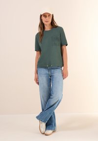 T-shirt en coton vert avec une poche texturée, manches courtes et un ourlet à cordon de serrage, associé à un jean flare bleu clair et des baskets blanches.