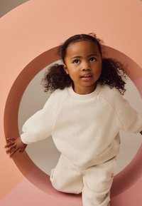 Ensemble sweat-shirt et pantalon couleur crème tachetés porté par un enfant, positionné sur un fond circulaire rose. La tenue présente des poignets et un ourlet côtelés.