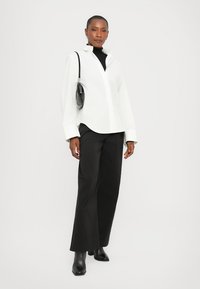 Chemise blanche à manches surdimensionnées, associée à un pantalon large noir et des bottines noires ; petit sac à main noir en accent.