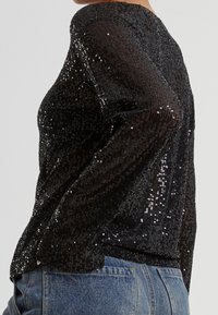 Haut noir à manches longues recouvert de sequins brillants, porté avec un jean en denim bleu, vu de profil du milieu du torse jusqu'au cou.