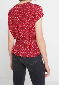 Červený top s krátkým rukávem a peplum lemem, s texturovaným vzorem bílých a modrých hvězd, nošený přes tmavě šedé džíny.