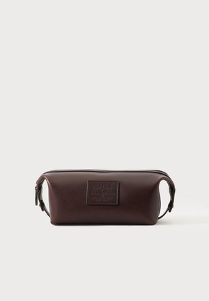 Trousse de toilette rectangulaire en cuir marron avec écusson de marque en relief sur le devant et poignée latérale sur fond blanc.