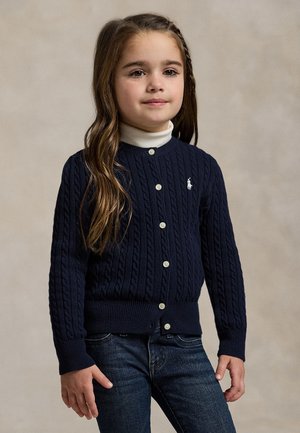 Jeune fille aux longs cheveux bruns portant un cardigan bleu marine en maille torsadée par-dessus un col roulé blanc et un jean foncé, debout devant un fond uni.