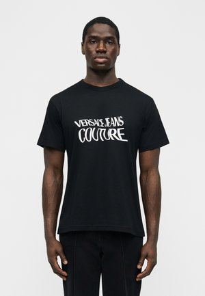 Modèle masculin portant un T-shirt noir avec le texte blanc « Versace Jeans Couture », debout devant un fond uni clair.