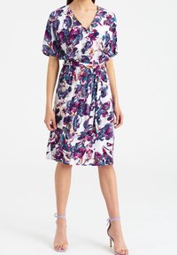 Sukienka w kwiatowy wzór wykonana z lekkiego materiału, z dekoltem w serek, krótkimi rękawami i talią podkreśloną paskiem. Paleta kolorów obejmuje fioletowy i biały.