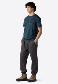 Teal kortærmet t-shirt med sort logo-patch, parret med grå joggingbukser og brune vandrestøvler med sorte såler. Tøjet har en afslappet pasform.