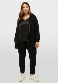 Felpa nera con zip e colletto a V in satin, jeans neri e sneakers bianche. Tessuto morbido, vestibilità rilassata e dettagli con cappuccio regolabile.