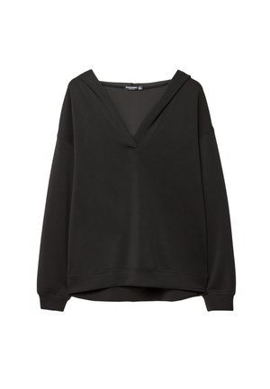 Kobieta w czarnym topie z dekoltem w serek i legginsach trzyma pikowaną czarną torebkę na ramię, ma na sobie białe skarpetki w paski i czarne tenisówki, brązowe tło.