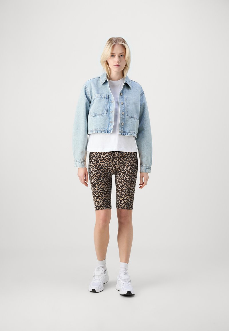 Giacca di jeans blu chiaro corta sopra una t-shirt bianca, abbinata a pantaloni corti da ciclismo con stampa leopardata marrone e sneakers bianche. Design semplice e vestibilità casual.