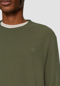 Olivgrüner, gerippter, langärmeliger Pullover mit Rundhalsausschnitt, der ein kleines erhabenes Logo auf der Brust und einen strukturierten Stoff aufweist.