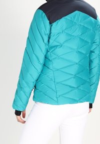 Turkusowa kurtka puchowa z diamentowym pikowaniem, czarnymi ramionami i elastycznymi mankietami. Zaprojektowana z myślą o cieple, o gładkiej, wyściełanej fakturze.