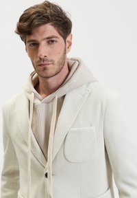 Blazer bianco con texture, colletto a revers e una tasca, indossato sopra un hoodie beige a costine con coulisse.