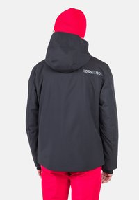 Giacca nera impermeabile con cappuccio e caratteristiche regolabili, con il logo "Rossignol" in grigio, abbinata a pantaloni di un rosso brillante.