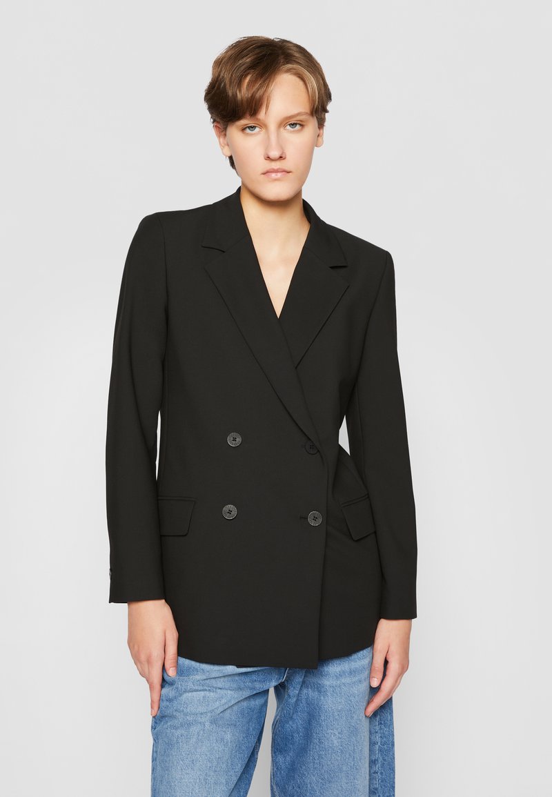 Blazer nero doppiopetto con rever a punta, dotato di due tasche a pattina e bottoni neri. Indossato sopra pantaloni in denim azzurri chiari.