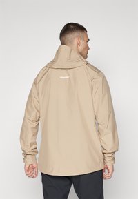 Beige vattentät jacka med hög krage, justerbar huva och långa ärmar. Har en tryckt logotyp på baksidan och en strömlinjeformad design.