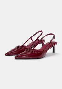 Talons slingbacks pointus bordeaux brillants avec des fines sandales et un petit talon kitten sur un fond blanc.