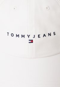 Valge puuvillane pesapalli müts, millel on tumesinises broderingus "TOMMYJEANS" ning punane, valge ja sinine embleem. Tugevdatud kuju ja nähtav õmblemine.