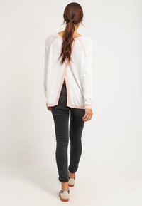 Donna con capelli lunghi raccolti in una coda di cavallo, che indossa un top bianco a maniche lunghe con uno spacco sulla schiena, jeans skinny neri e sneaker bianche, mentre si allontana.