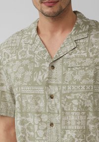 Korte mouw, groene Hawaiiaanse shirt met witte bloem- en geometrische patronen, knoopsluiting en een zak aan de linkerkant op de borst.