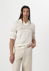 Pull en laine crème avec des rayures horizontales, doté de manches courtes et d'un col en V. Associé à un pantalon beige clair ajusté.