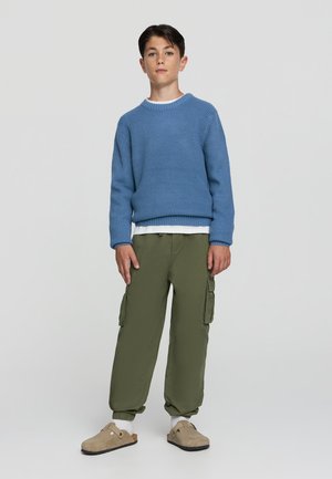Maglione blu lavorato a maglia, pantaloni cargo verdi con tasche laterali, zoccoli beige e calzini bianchi. In piedi contro uno sfondo neutro.