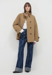 Donna in piedi contro uno sfondo bianco che indossa una giacca beige con bottoni, una maglietta bianca, jeans larghi a righe blu e scarpe nere con punta.