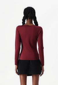Haut bordeaux à manches longues avec un design ajusté, texture lisse et col rond, associé à une jupe noire. Vue arrière présentée.