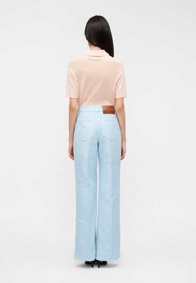 Φαρδιά παντελόνια σε ανοιχτό μπλε με απαλό ύφασμα, διακοσμημένα με ένα καφέ δερμάτινο ταμπελάκι στο πίσω μέρος. Συνδυάζονται με ένα ροζ πουκάμισο με γιακά.