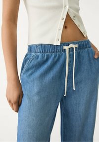 Kvinna klädd i ljusblå denim joggerbyxor med vit dragsko och vit ribbad ärmlös top med knappar, händerna avslappnade vid sidorna.