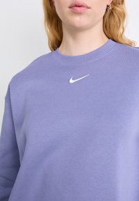 Sudadera de cuello redondo morada, hecha de una mezcla de algodón, con el logo blanco de Nike en el pecho. Textura suave y cuello acanalado.