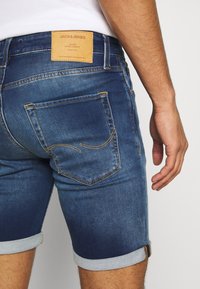 Muž v modrých ohrnutých denimových šortkách s hnědou koženou cedulkou Jack & Jones na pásku, částečně viditelná bílá košile.