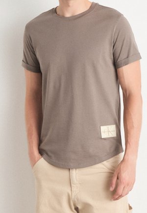 Braunes T-Shirt mit kurzen Ärmeln und Rundhalsausschnitt aus weichem Stoff, mit einem rechteckigen Logopatch auf der unteren rechten Seite.