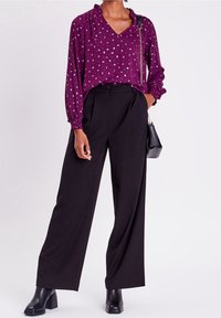 Blouse violette à pois blancs, décolleté en v et manches froncées, associée à un pantalon large noir taille haute et des bottes à talons épais noires.