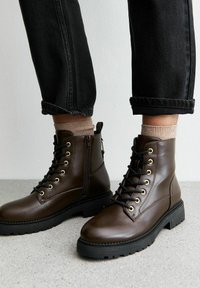 Bottes de cheville en cuir marron avec œillets dorés, semelles en caoutchouc noires et fermeture éclair sur le côté. Portées avec un pantalon en jean gris à revers et des chaussettes marron clair.
