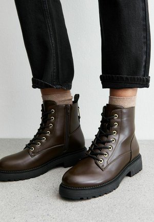 Botas de tobillo de cuero marrón con ojales dorados, suelas de goma negras y una cremallera lateral. Llevadas con jeans gris con puños y calcetines marrones claros.