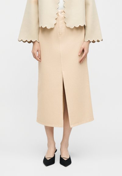 Γυναίκα με μπεζ midi φούστα με εμπρόσθια σχισμή, σακάκι με κυματιστά άκρα και μαύρα μπαλαρίνες με λεπτό λουράκι, στέκεται μπροστά σε λευκό φόντο.