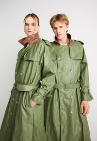 Διπλής σειράς παλτά τρεντς από δέρμα σε ελιά πράσινο με καφέ διακοσμητικές λεπτομέρειες στον γιακά, ζώνη στη μέση και μεγάλα κουμπιά, με λεία υφή.