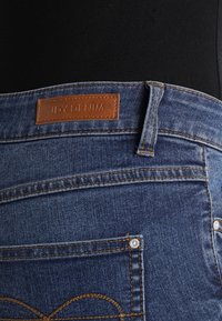 Jeans de mezclilla azul con costuras marrones y una etiqueta de cuero con la inscripción "JDY DENIM" que se llevan sobre una camiseta negra.