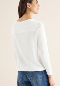 Haut blanc à manches longues avec un décolleté incurvé et un dos semi-transparent, associé à un jean en denim bleu. Texture douce et lisse.