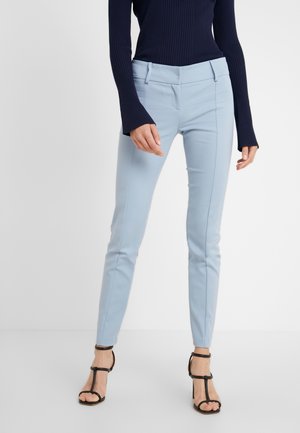 Mujer que lleva pantalones ajustados azul claro y sandalias negras de tacón alto con tiras, de pie frente a un fondo liso.