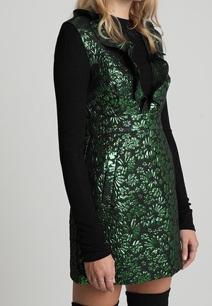 Grøn metallisk blomstre brokade ærmeløs kjole med flæsekrave båret over en sort langærmet top og sorte støvler til låret.