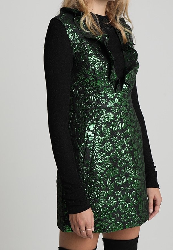 Robe sans manches en brocart floral métallique vert avec un col volanté portée sur un haut noir à manches longues et des bottes noires montantes.