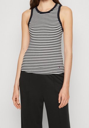 Kobieta w czarno-białym poziomo paskowanym bezrękawniku z czarną lamówką, w połączeniu z czarnymi spodniami na jasnoszarym tle.