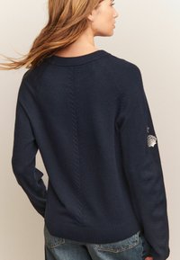 Marinblå tröja i mjuk stickad kvalitet med rund halsringning, ribbade ärmslut och broderad motiv av snäcka på vänster ärm.