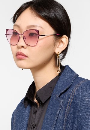 Giovane donna con occhiali da sole rosa oversize, orecchini a catena d'oro, camicia scura e giacca di jeans blu, che guarda di lato su sfondo bianco.