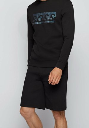 Mies, joka pukeutunut mustaan pitkähihaiseen collegepaitaan, jossa sininen "BOSS"-teksti, ja mustiin shortseihin, valkoista taustaa vasten.